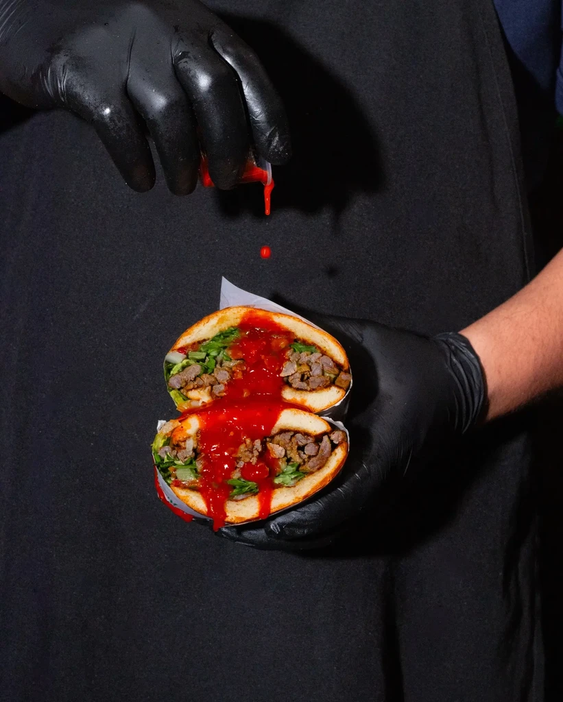 Gloved hands hold a sandwich with meat, greens, and dripping red sauce.