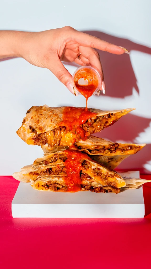 Hand drizzles red sauce over two stacked, meat and cheese quesadillas.