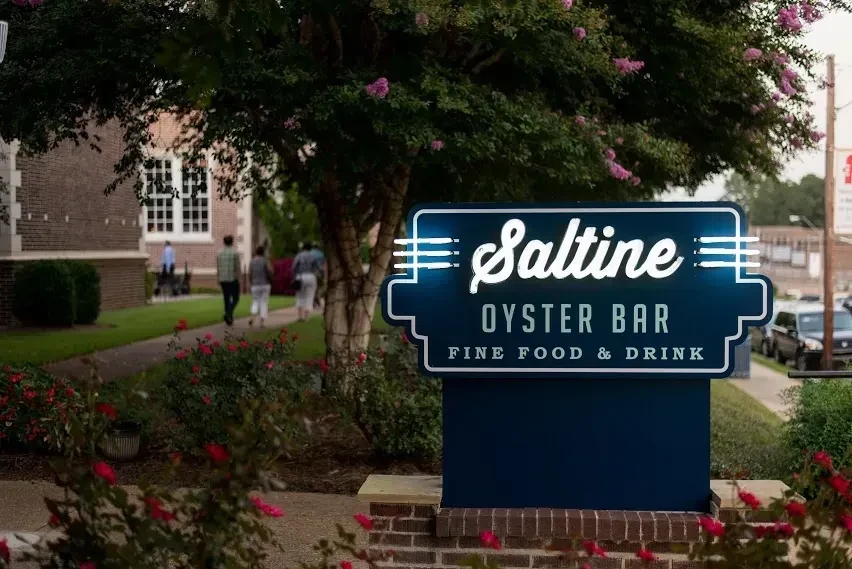 Blue neon "Saltine Oyster Bar" sign illuminated among outdoor plants.
