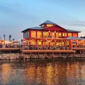 Red waterfront restaurant building at dusk with illuminated patios and people.