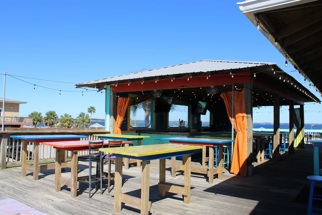 Outdoor deck bar with colorful tables, water, and palm trees.