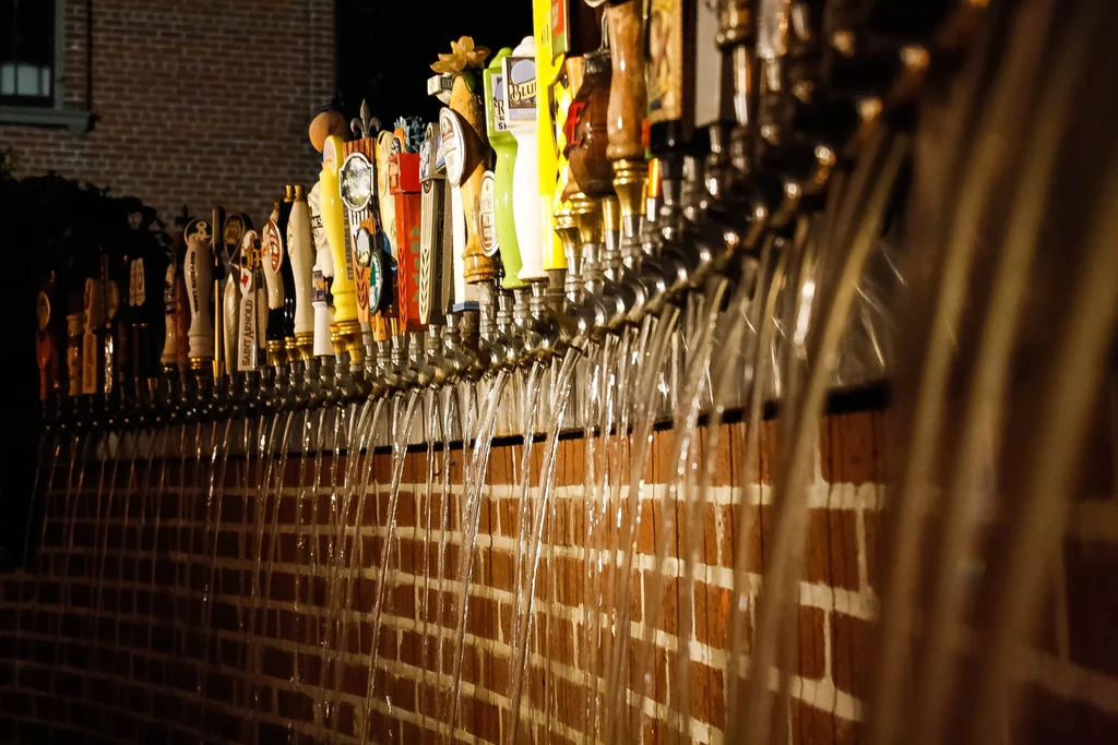 Long row of beer taps pouring beer in front of a brick wall.