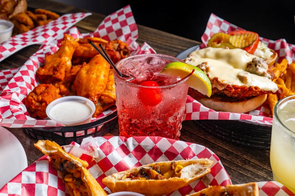Red cocktail, chicken wings, burger, and pastry on a dark table.