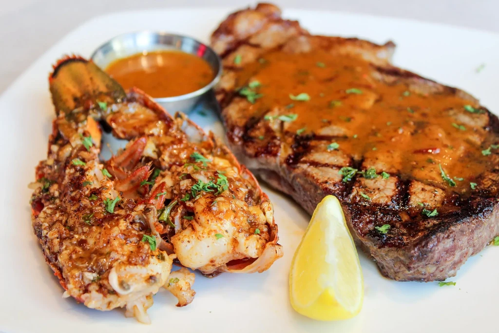 Plated grilled steak, butterflied lobster tail, lemon, and dipping sauce.