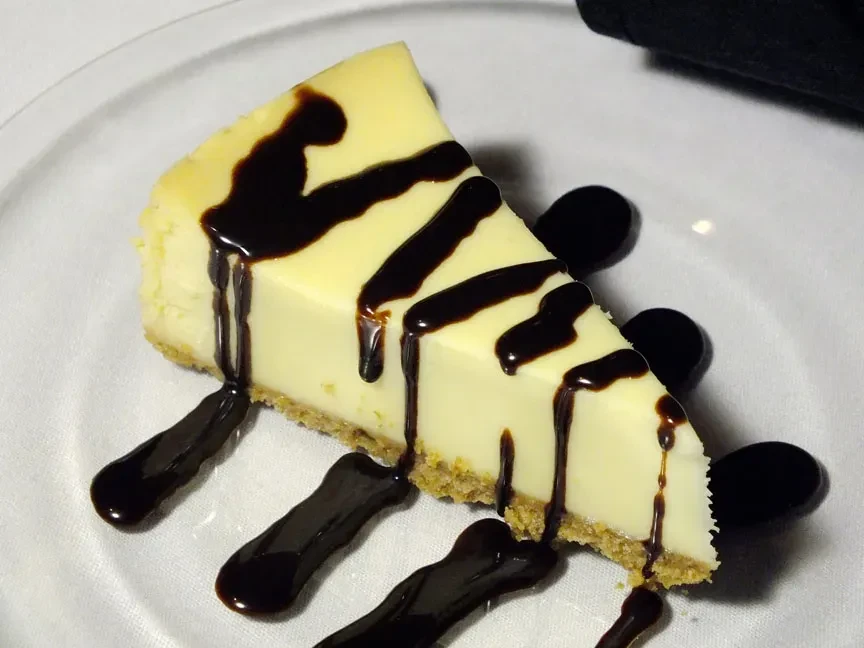 Cheesecake slice with graham cracker crust and chocolate syrup drizzle.