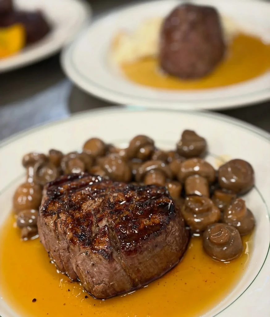 Grilled meat with sauce and mushrooms on plate, blurred meal behind.