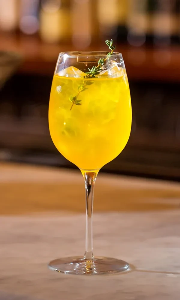 Yellow drink in tulip glass with ice and thyme garnish.