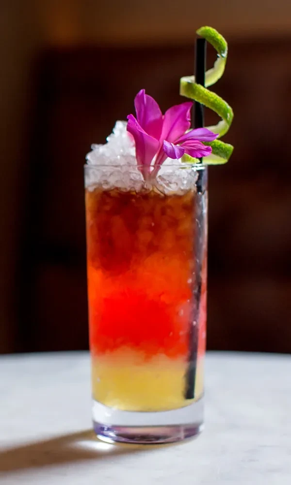 Three-layered cocktail with crushed ice, tropical flower, and lime zest.