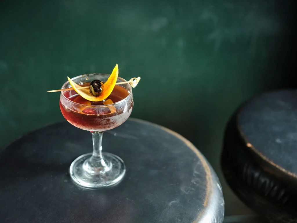 Chilled reddish-brown cocktail in coupe glass, garnished with orange and cherry.