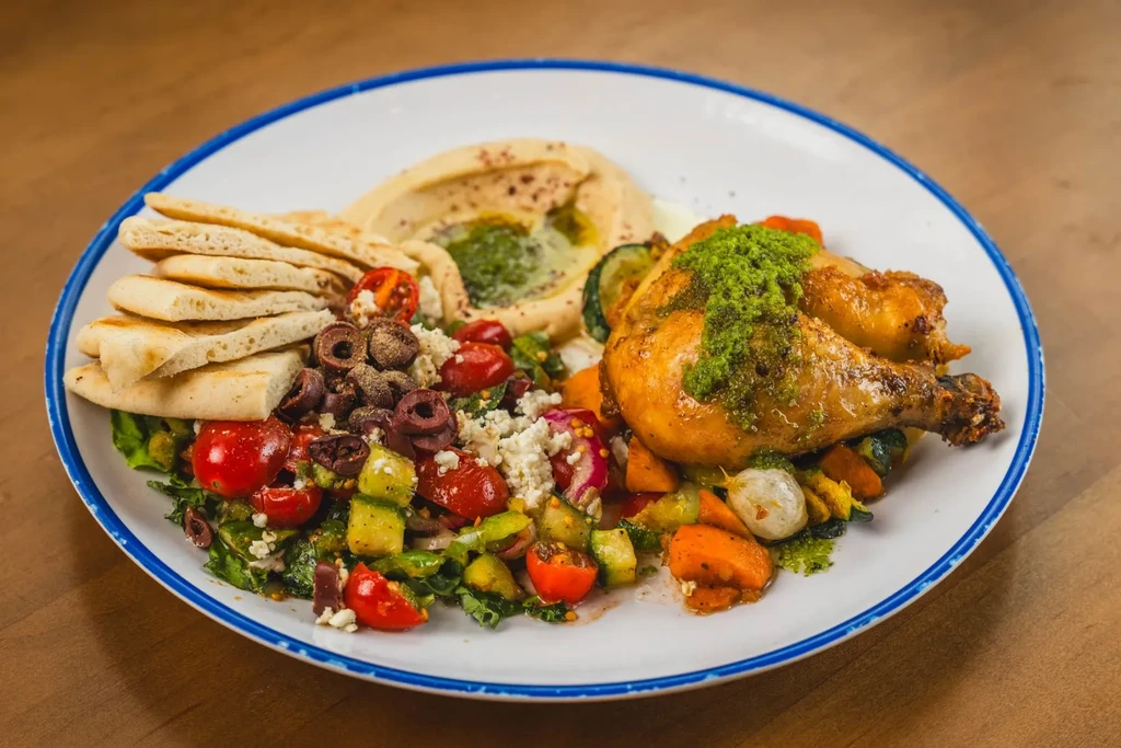 Roasted chicken, hummus, pita, fresh salad, and roasted vegetables plated.