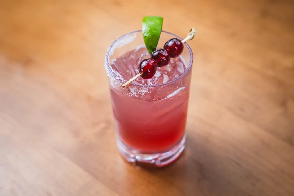 Reddish-pink cocktail with ice, berry skewer, lime wedge, and frosted rim.