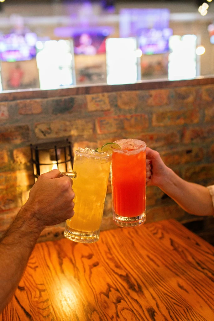 Close-up of two hands toasting vibrant iced drinks at a bar.
