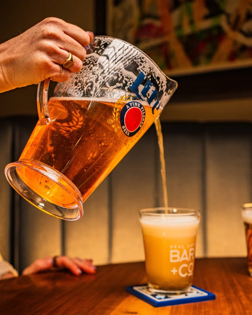 Person pours "A FINE PILSNER BEER" into a "BAR + CO" glass.