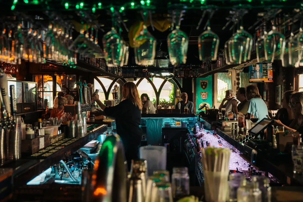 Bartender serves patrons at a brightly lit bar with hanging wine glasses.