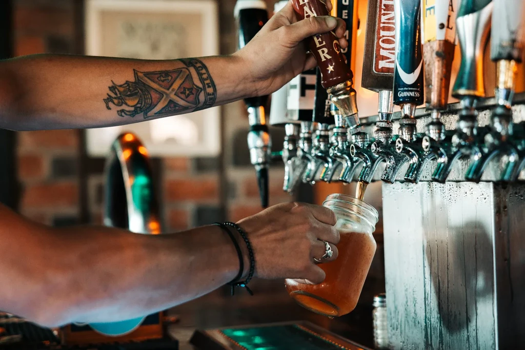 Tattooed arm pours amber beer from a tap into a glass.