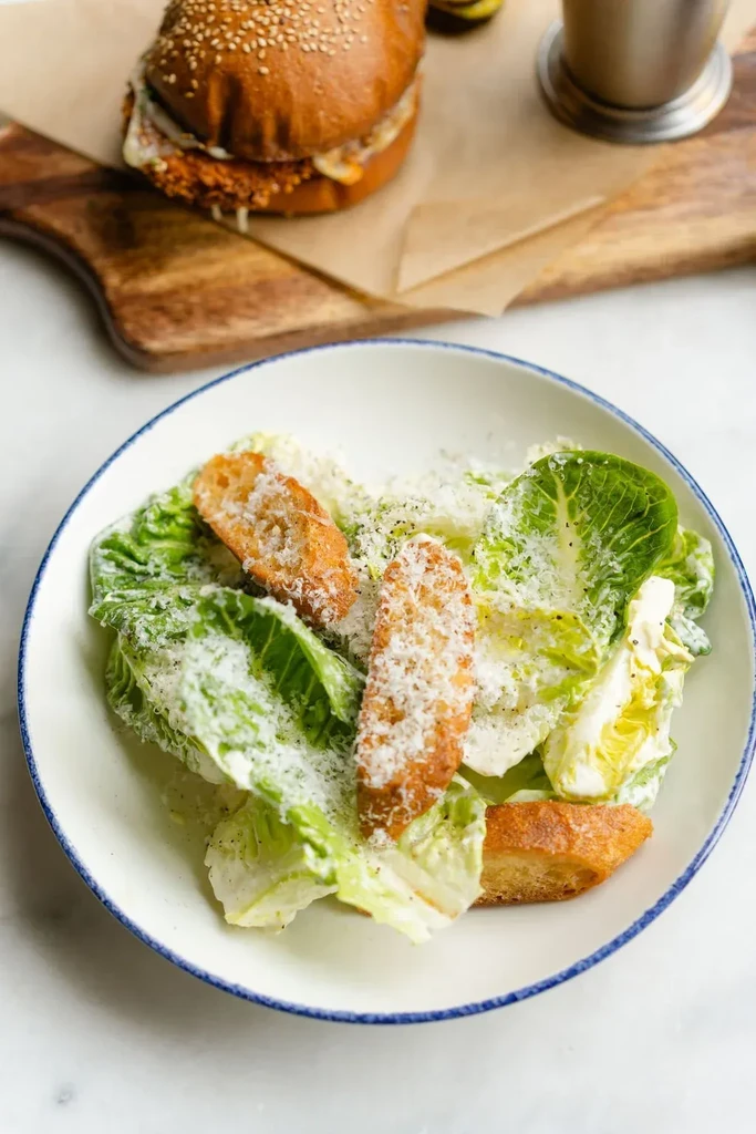 Close-up of Caesar salad bowl, with blurred burger in background.