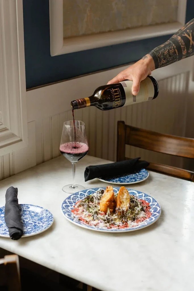Hand pouring Quilt Napa Valley red wine beside a meat dish.