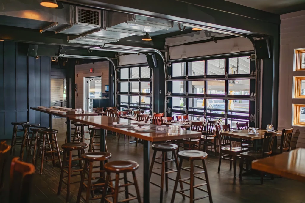 Modern restaurant interior with communal table, industrial elements, and large windows.