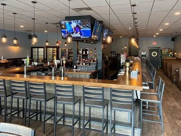Empty modern bar with L-shaped counter, liquor display, and sports TVs.
