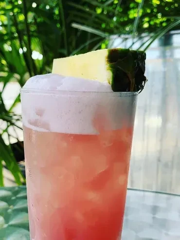 Vibrant pink-red drink with foam, pineapple garnish, lush green background.