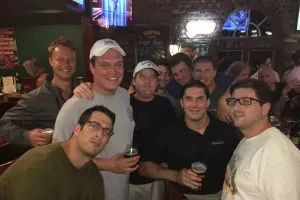 Approximately nine men smile, holding drinks in a casual bar.