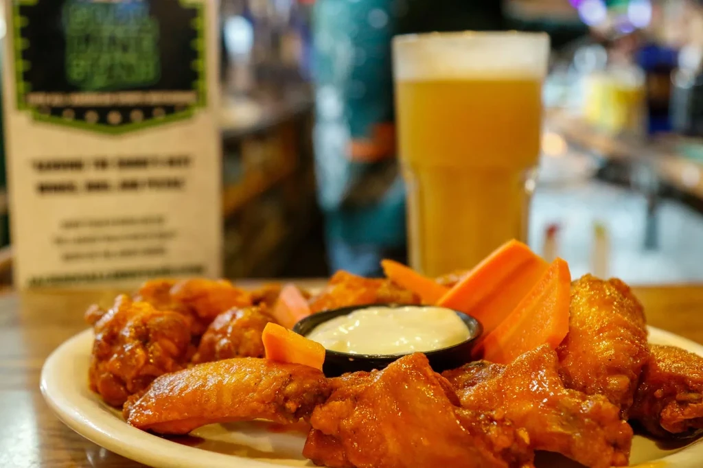 Close-up of buffalo chicken wings, carrots, white dip, and blurred beer.