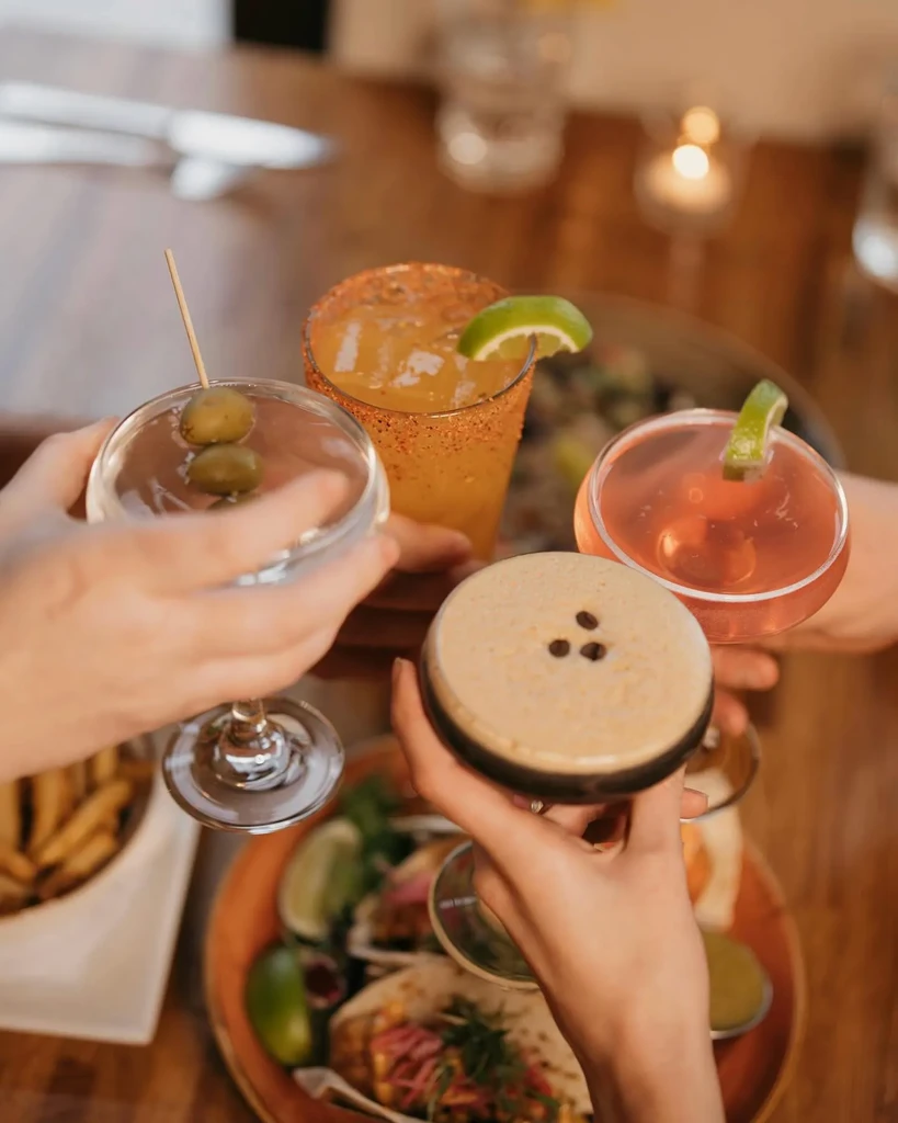 Four hands toast diverse cocktails over a spread of tacos and fries.
