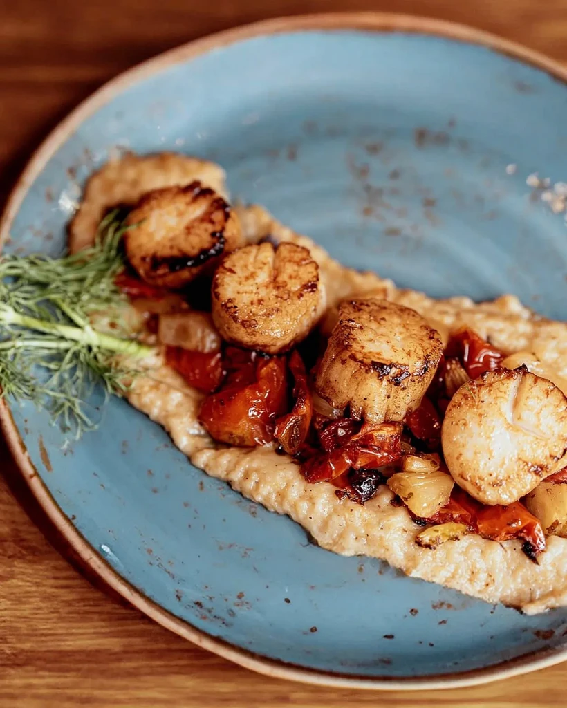 Seared scallops on blue plate with puree, roasted vegetables, and dill.