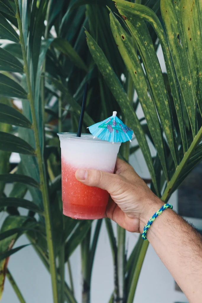 Hand holds layered frozen drink, straw, umbrella, against palm leaves.