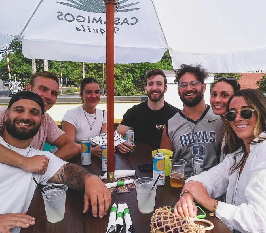 Seven young adults smile at an outdoor table with various drinks.