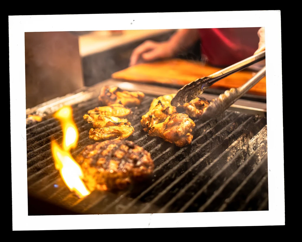 Hands with tongs grill chicken wings and a burger over open flames.