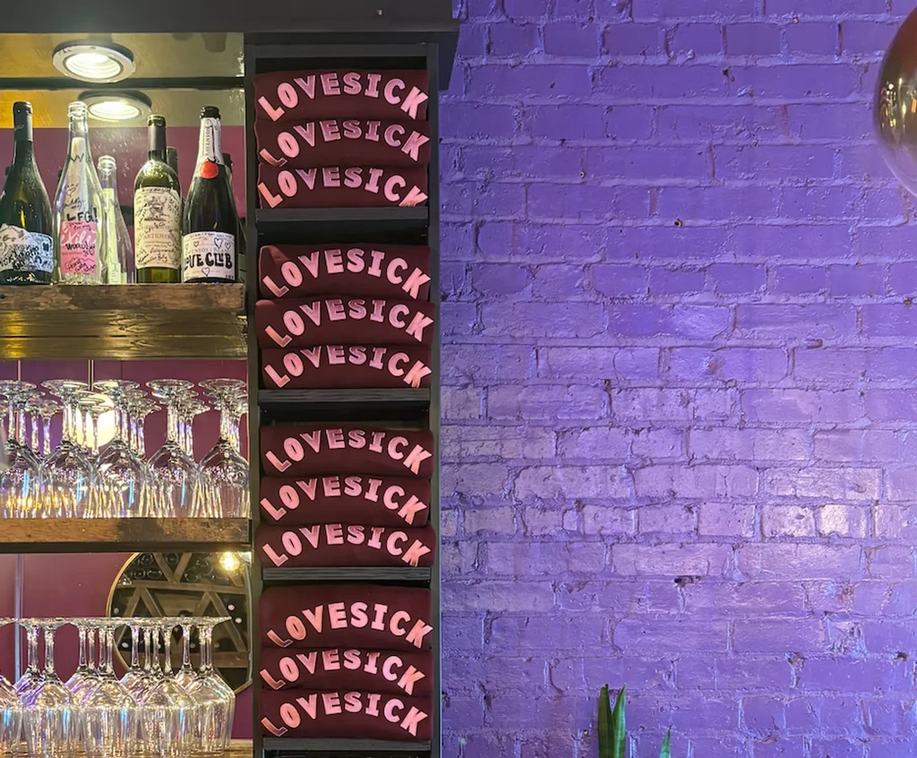 Bar interior with bottles, glasses, LOVESICK shirts, and purple wall.
