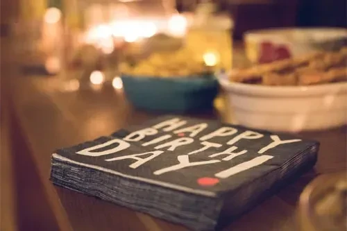 Stack of black "Happy Birthday!" napkins on wood, blurred festive background.