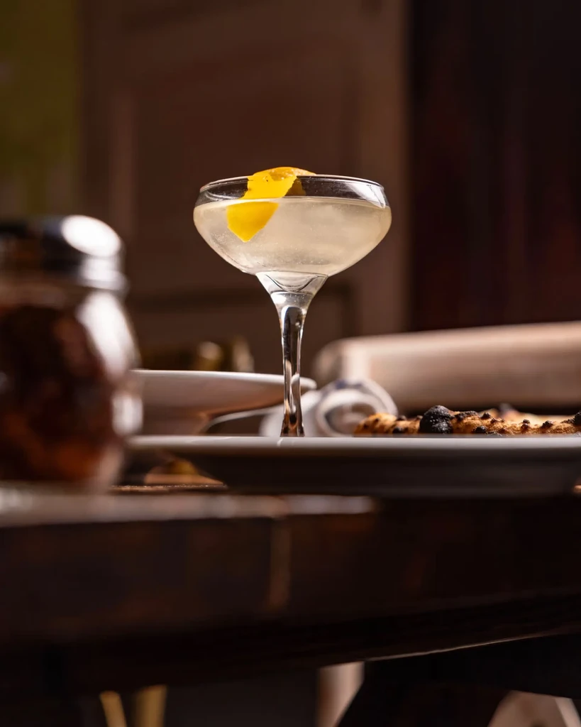 Light cocktail in coupe glass with lemon, blurred bar background.