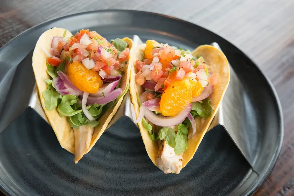 Two tacos with protein, greens, orange, salsa in a metal holder.