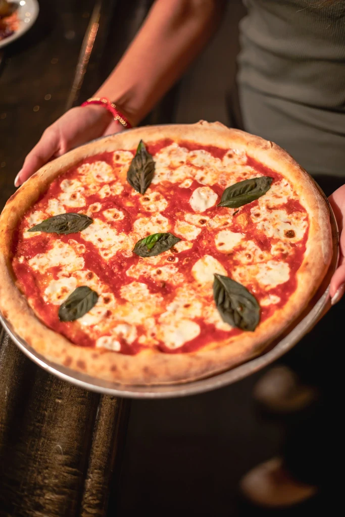 Warmly lit Margherita pizza held by hands on a metal pan.