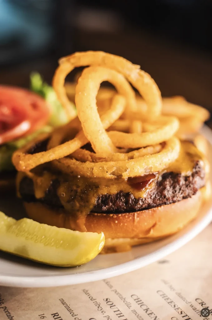 A cheeseburger piled with onion rings, melted cheese, lettuce, tomato, pickle.