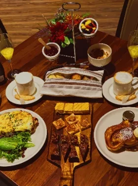 Warmly lit wooden table laden with diverse brunch foods, drinks, and flowers.
