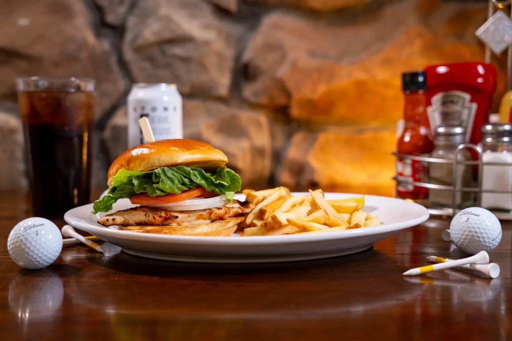 Close-up: grilled chicken sandwich, fries, drinks, golf balls on wooden table.