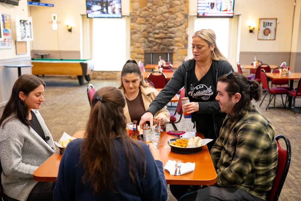 Server at BAR Baddyshack Grill serves drinks to patrons at a table.