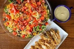 Colorful salad, fried chicken strips, and dressing on a wooden table.