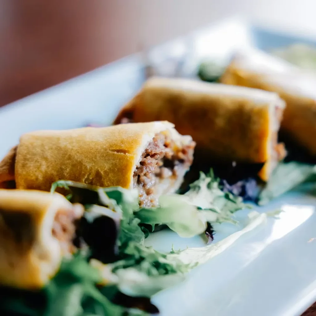 Golden-brown spring rolls with minced meat filling and greens on a white plate.