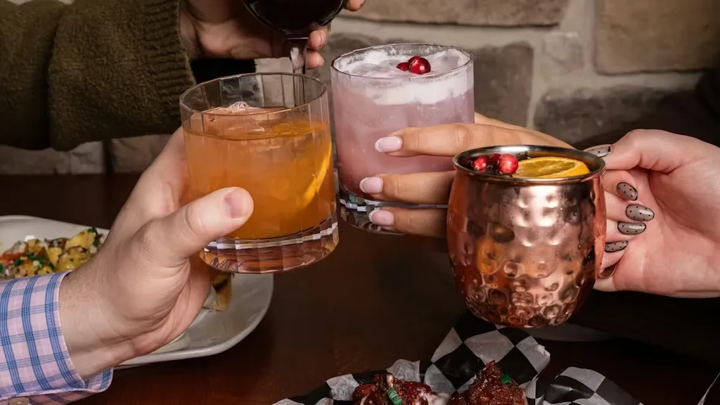 Four hands toast diverse cocktails and appetizers in a cozy setting.