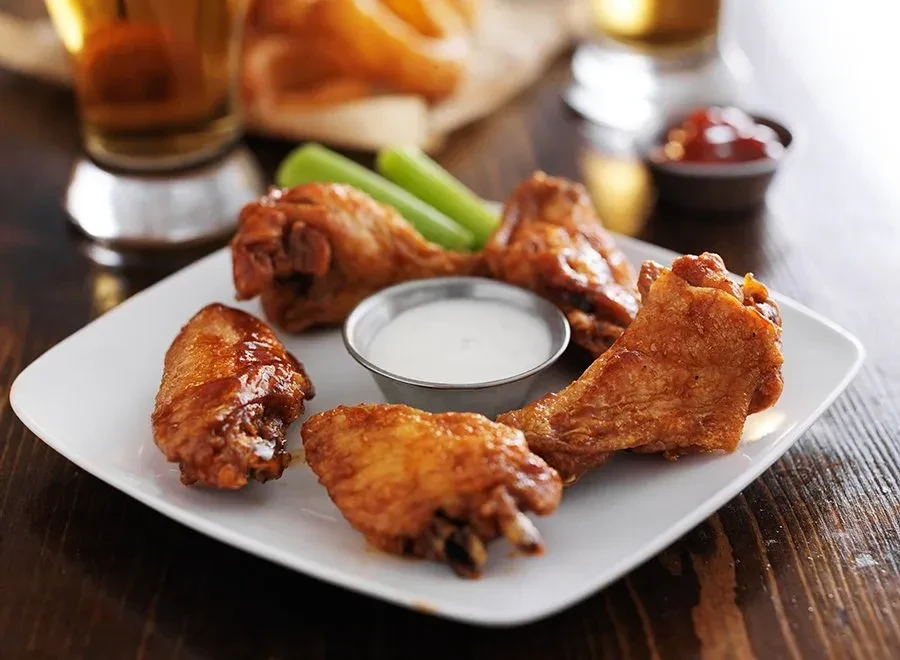 Five crispy glazed chicken wings, white dip, celery, and drinks on wood.