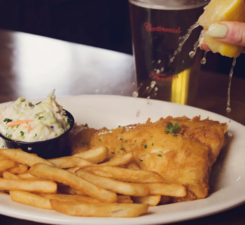 Hand squeezing lemon over golden fish, crispy chips, and coleslaw.