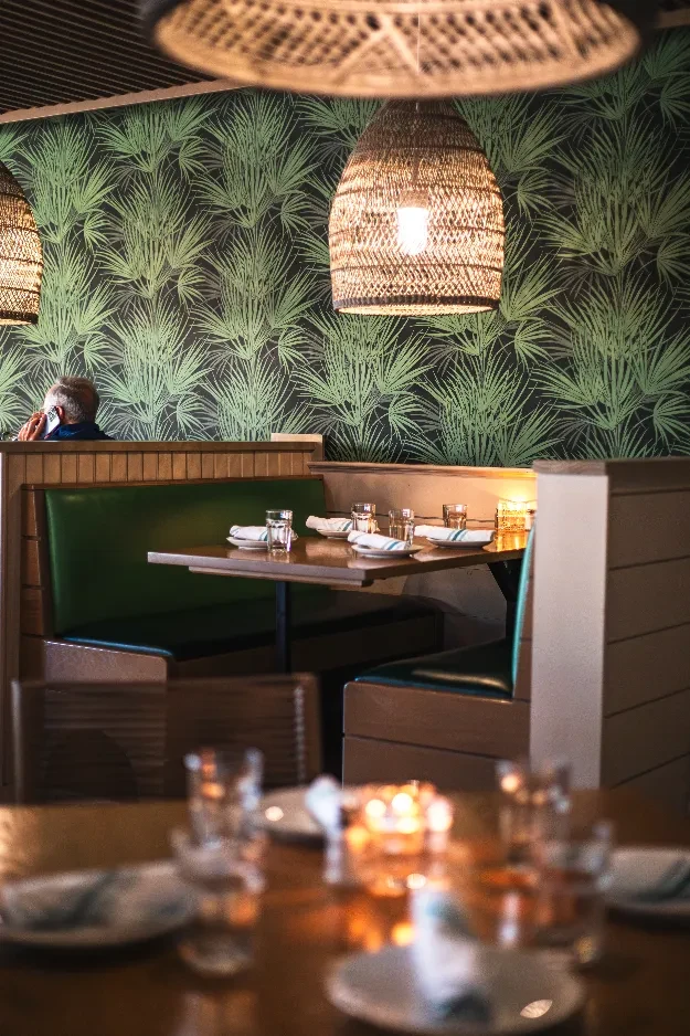 Restaurant interior with green palm leaf wallpaper, woven lights, dark green booths.