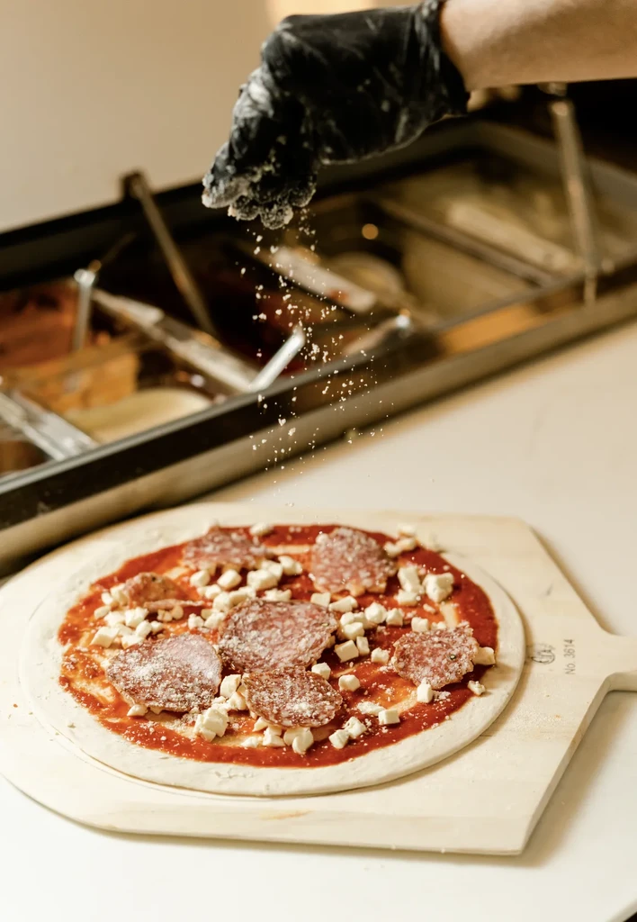 Gloved hand sprinkles powder on raw pepperoni pizza with cheese.