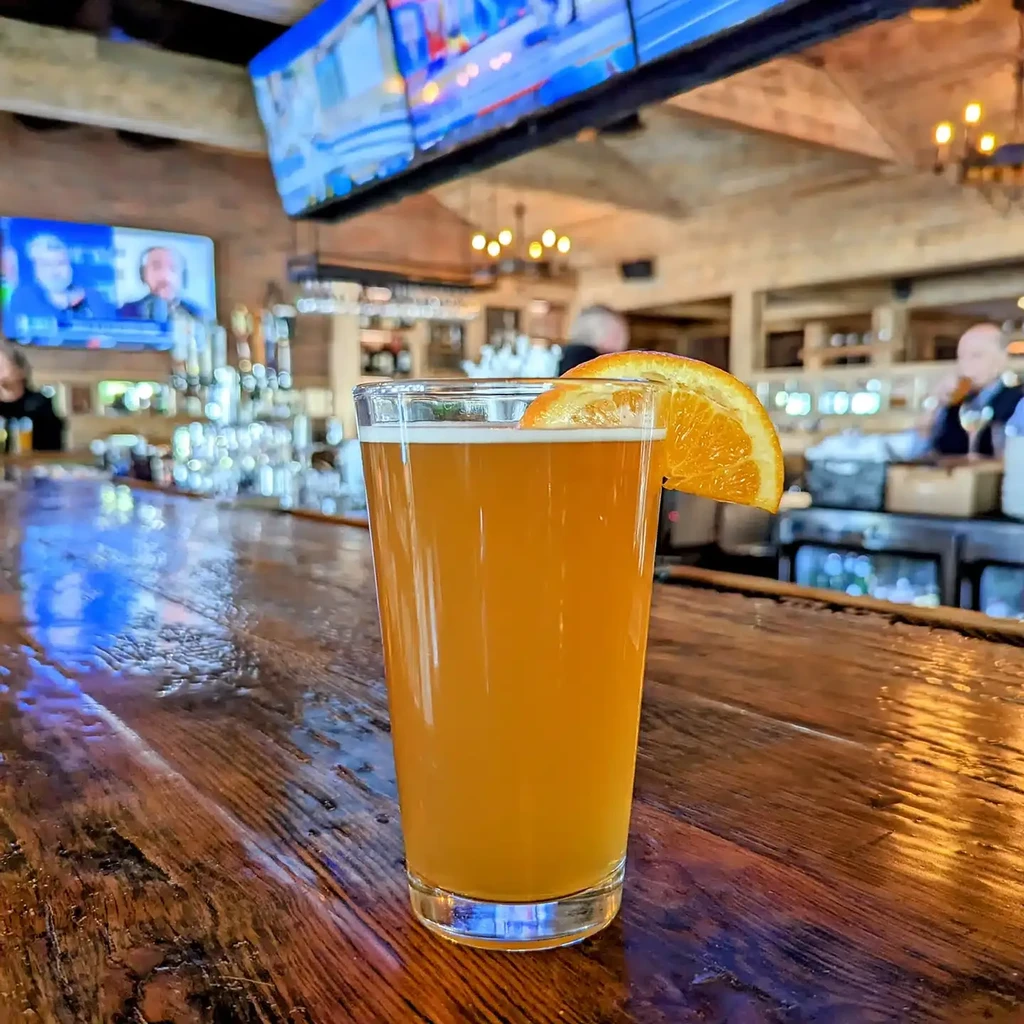 Amber drink with orange slice on wooden bar in a lively pub.