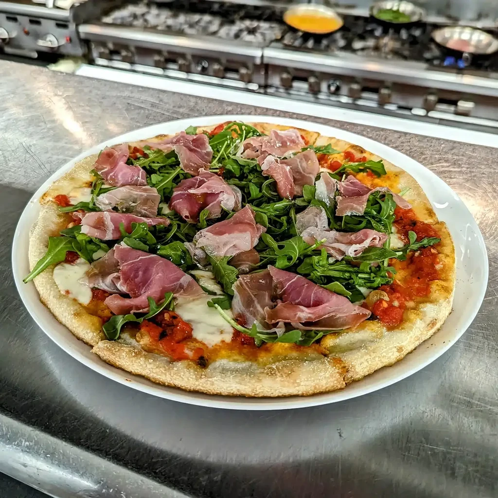 Pizza with tomato sauce, mozzarella, arugula, and prosciutto in a kitchen.