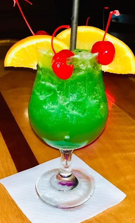 Green slushy cocktail in stemmed glass with cherries and orange slices.